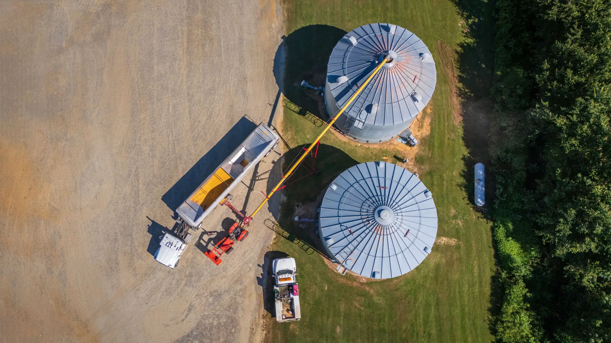 Top-down view of a grain trailer unloading into an auger beside two storage bins